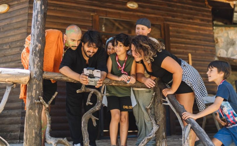 A diverse group of people joyfully watching drone footage on a wooden cabin porch.